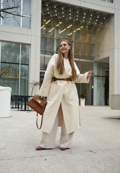Mujer con un abrigo largo color crema y pantalones beige, sosteniendo un bolso marrón, de pie frente a un edificio moderno de vidrio, sonriendo y mirando hacia un lado.