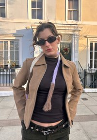 Veste en cuir couleur fauve avec une fermeture éclair, col roulé noir et écharpe texturée aux couleurs pastel. Lunettes de soleil noires et boucles d'oreilles anneaux.