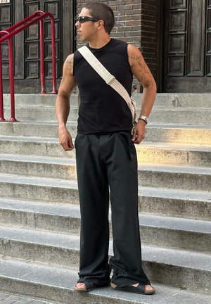 Hombre con tatuajes que lleva gafas de sol, camiseta negra sin mangas, pantalones negros, sandalias y bolso cruzado, de pie en escalones de piedra al aire libre.