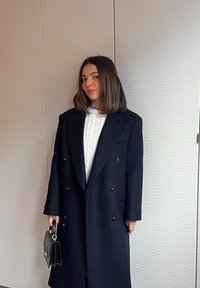 Γυναίκα με καστανά μαλλιά μέχρι τους ώμους φορά μακρύ, μαύρο παλτό διπλής σειράς κουμπιών πάνω από ένα λευκό πουλόβερ με πλεκτό σχέδιο, κρατώντας μια μαύρη τσάντα.