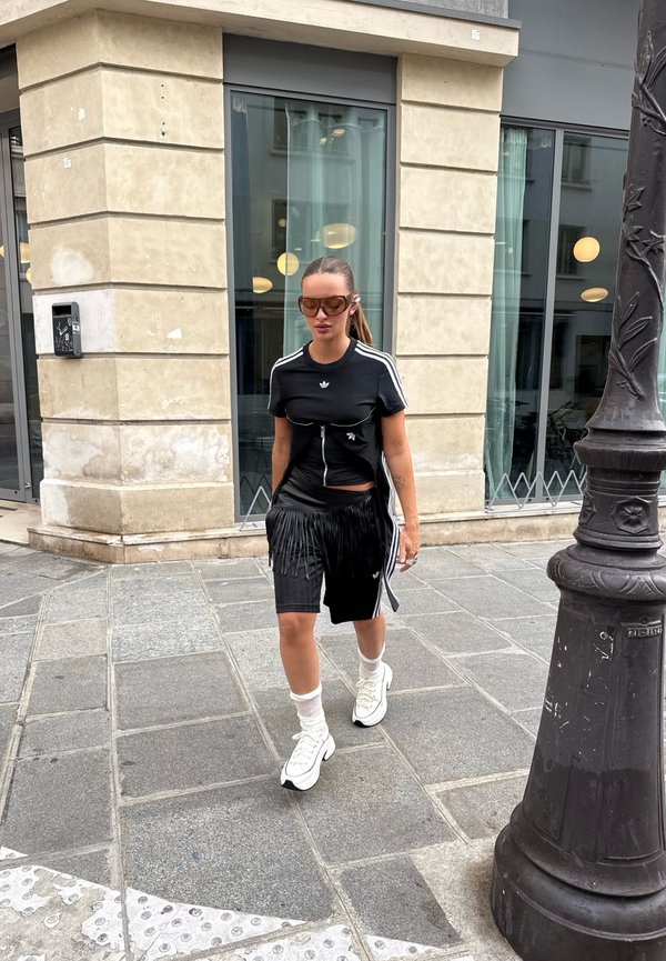 Mujer vestida con ropa deportiva negra de Adidas, zapatillas blancas y gafas de sol grandes caminando por una acera de la ciudad cerca de un edificio de piedra y una farola.