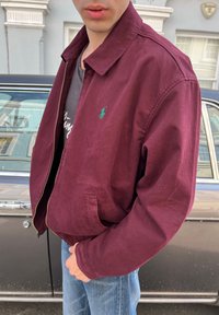 Giovane uomo con giacca zip bordeaux e jeans blu in piedi accanto a un'auto d'epoca scura su una strada di città.