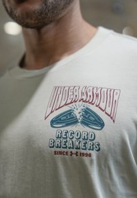 T-shirt de manga curta cor creme claro com impressão gráfica de dois pares de ténis de corrida, rotulados "Under Armour Record Breakers" em vermelho e azul.