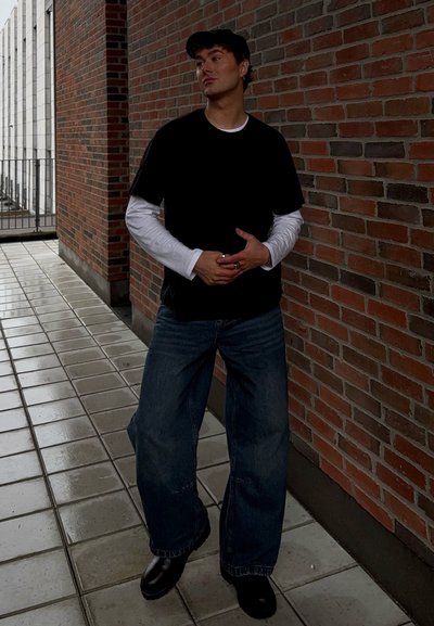 Camiseta negra de manga corta sobre una camiseta blanca de manga larga, jeans de mezclilla sueltos y botas negras. Fondo de pared de ladrillos y suelo de azulejos.