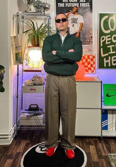 Sudadera verde con cremallera, pantalones de vestir beige y zapatos rojos. Los accesorios incluyen gafas de sol. El fondo presenta estanterías con decoración y zapatillas.