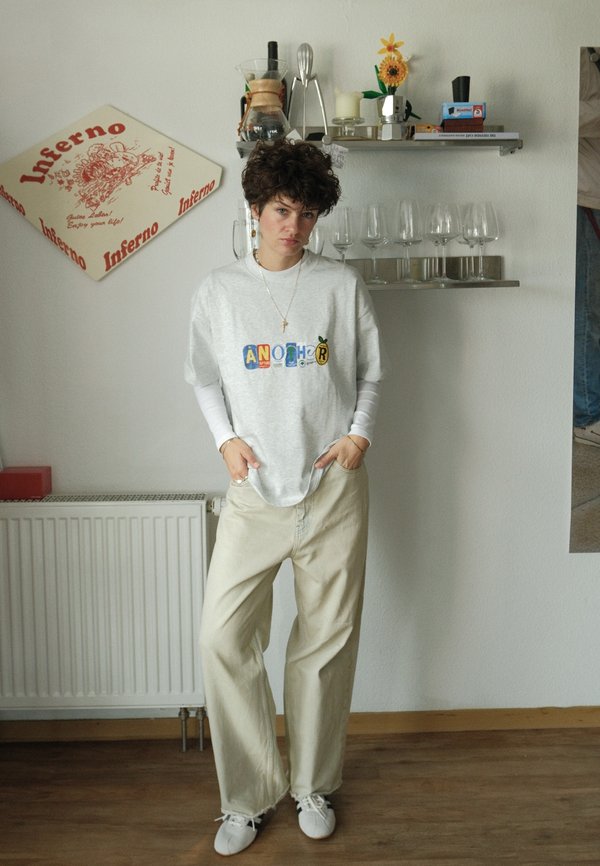 Una persona joven con una sudadera blanca con un diseño estampado y unos pantalones beige holgados está de pie en una cocina, con cristalería y utensilios en las estanterías detrás.