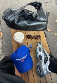 Sac en cuir noir, casquette Polo Sport bleue, baskets argentées, lunettes de soleil noires, gobelet à café jetable, bague bleue sur une table en bois en plein air.