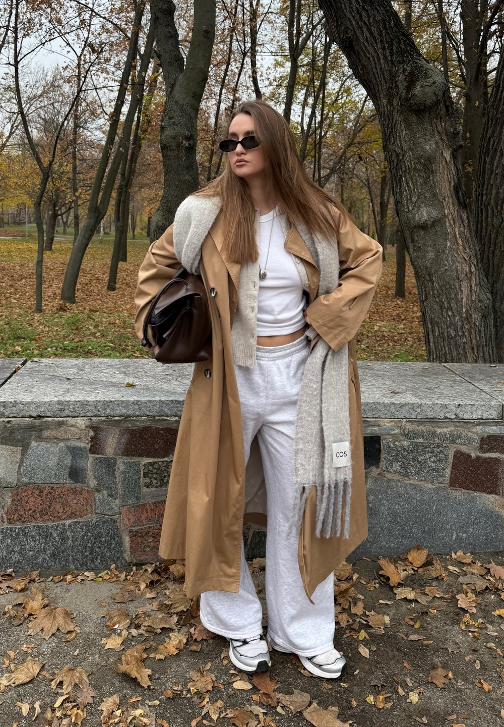 Tânără cu păr lung purtând un pardesiu bej, pantaloni albi de lounge, adidași, ochelari de soare negri și ținând o geantă maro într-un parc cu frunze de toamnă.