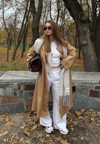 Tânără cu păr lung purtând un pardesiu bej, pantaloni albi de lounge, adidași, ochelari de soare negri și ținând o geantă maro într-un parc cu frunze de toamnă.