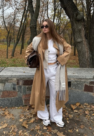 Joven con cabello largo que lleva un abrigo beige, pantalones cómodos blancos, zapatillas deportivas, gafas de sol negras y sostiene un bolso marrón en un parque con hojas de otoño.