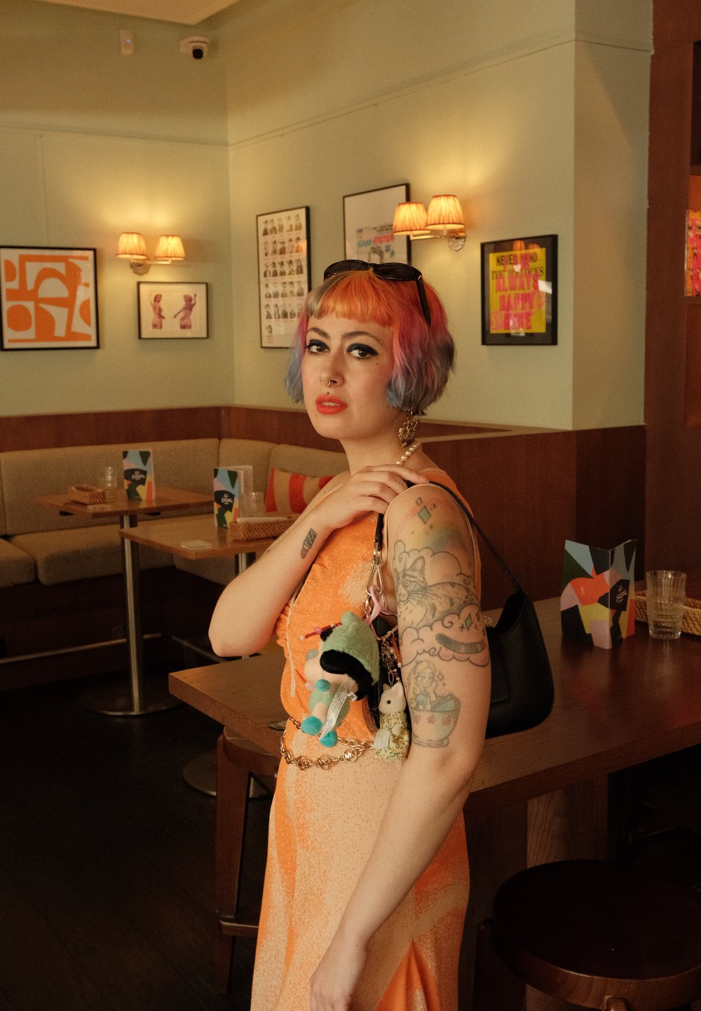 Een mouwloos oranje jurkje met een gradientontwerp, voorzien van een gouden riem. Zwarte tas met kleurrijke pluche sleutelhangers eraan. Tattoo's zichtbaar op de arm.