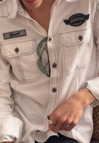 Personne portant une chemise boutonnée blanc cassé avec des coutures noires, des écussons indiquant "Brandt" et "Addicted", tenant des lunettes de soleil et portant une bague en forme d'étoile.
