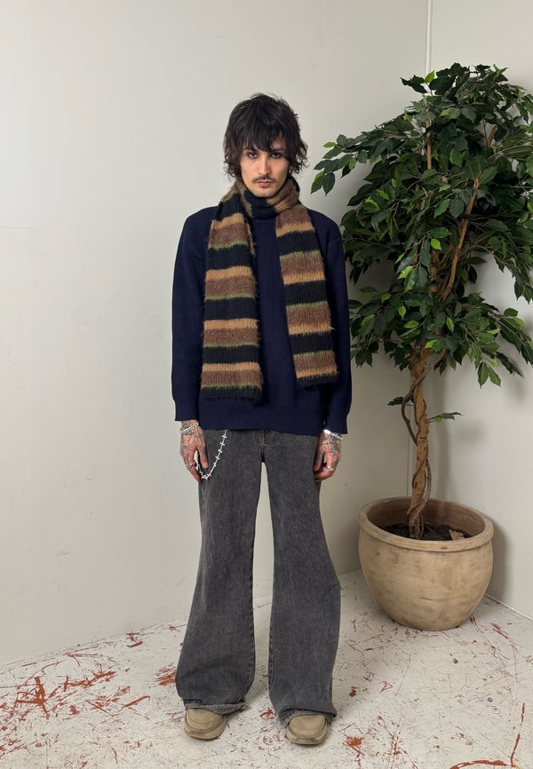 Mees seisab siseruumis, kandes triibulist pruuni ja musta sall, sinist kampsunit, halli laia jalaga pükse ja beeže kingi, kõrval roheline potitaim.