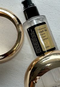 Bouteille transparente de COSRX Advanced Snail 96 Mucin Power Essence avec un capuchon noir, ornée d'une étiquette dorée et entourée de deux accents circulaires dorés brillants.