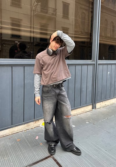 Persona que lleva una camiseta gris corta sobre una camiseta de manga larga gris, vaqueros negros anchos y desgastados, y zapatos negros, con auriculares alrededor del cuello.