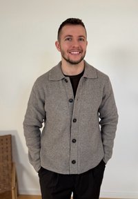 Χαμογελαστός άνδρας που στέκεται σε εσωτερικό χώρο, φορά γκρι πουλόβερ με κουμπιά και μαύρο παντελόνι, με τα χέρια στις τσέπες, μπροστά σε ένα απλό λευκό τοίχο.