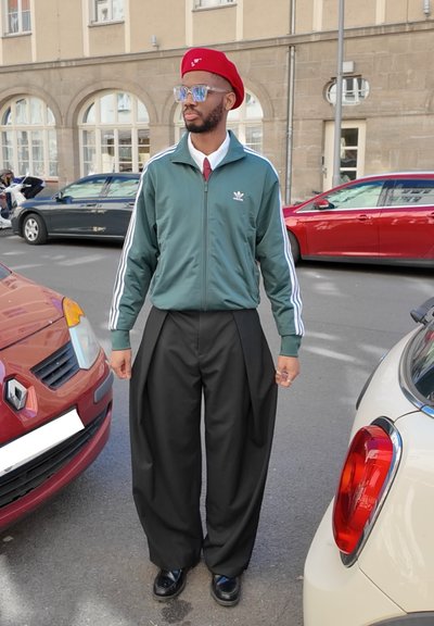 Chaqueta verde con cremallera y rayas blancas, pantalones negros holgados, boina roja y gafas, posando entre coches aparcados rojos y blancos.