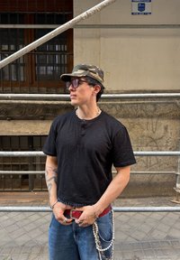 Fekete, rövid ujjú henley póló, kék laza farmernadrág, piros öv, ezüst lánc és terepmintás sapka átlátszó napszemüveggel; városi állványzat háttér.
