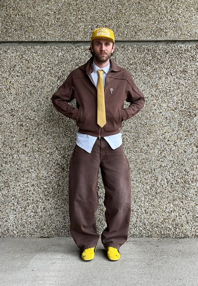 Chaqueta marrón con cremallera sobre una camisa blanca, corbata amarilla, pantalones marrones sueltos y zapatos amarillos. Gorra amarilla con texto. Fondo gris texturizado.