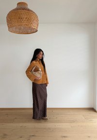 Femme en veste marron et jupe à pois tenant un sac beige, debout sur un sol en bois sous une grande lampe suspendue tissée, devant un mur blanc.