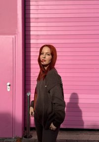 Νεαρή γυναίκα με κόκκινα μαλλιά που φοράει σκουρόχρωμο πουλόβερ, στέκεται μπροστά από ροζ παντζούρι και ροζ τοίχο κάτω από το φως του ήλιου.