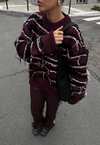 Pull en tricot maroon et noir avec des rayures texturées, coupe surdimensionnée, détails effilochés. Associé à un pantalon maroon et des chaussures noires à clous.