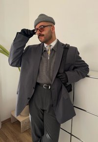 Harmaa räätälöity puku, jonka alla on vetoketjullinen villapaita, harmaa pipo, aurinkolasit ja mustat hanskat. Seisoo valkoisen kaapin vieressä, jossa on kasvi.