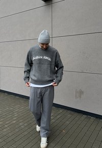 Grauer Strickpullover mit weißem Kursivtext, kombiniert mit hellgrauer, locker sitzender Hose und weißen Sneakers. Strukturierter Stoff, legerer Schnitt.