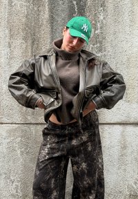 Osoba nosiaca zelenú čiapku, hnedý rolák, nadrozmernú tmavú koženú bundu a čierne nohavice posiate farbou, stojaca pri textúrovanej stene.