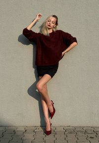 Suéter oversized cor vinho, saia mini preta e sapatos de salto alto vermelhos brilhantes. Modelo posa contra uma parede cinza texturizada, com uma postura casual.