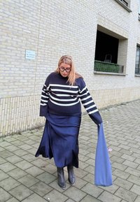 Μαύρο και άσπρο ριγέ πλεκτό πουλόβερ, σκούρα σατέν φούστα, μαύρες μποτάκια μέχρι τον αστράγαλο, στέκεται σε πλακόστρωτο δρόμο με ανοιχτόχρωμο τοίχο από πίσω.