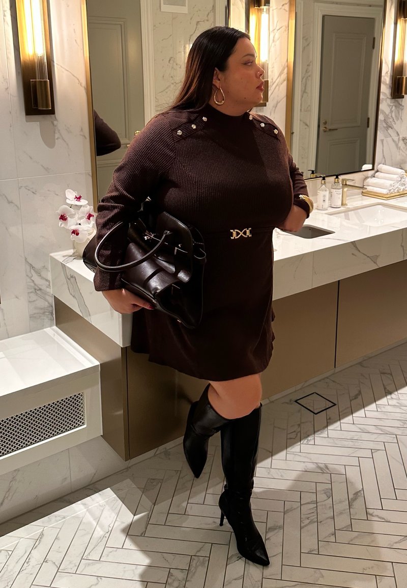 Brown ribbed sweater dress with gold accents, paired with black knee-high pointed boots and a black handbag, set against a marble backdrop.