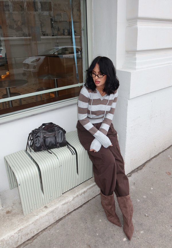 Femme aux cheveux noirs et lunettes assise sur un banc devant un bâtiment, portant un pull rayé, un pantalon marron et des bottes marron, à côté d'un sac à main noir.