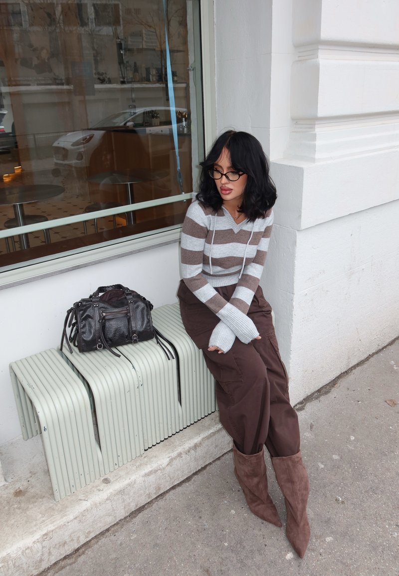Femme aux cheveux noirs et lunettes assise sur un banc devant un bâtiment, portant un pull rayé, un pantalon marron et des bottes marron, à côté d'un sac à main noir.