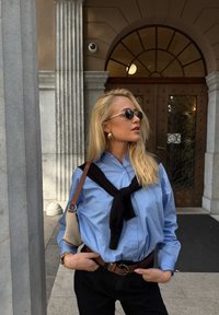 Camicia azzurro chiaro con maniche lunghe, un maglione nero legato attorno al collo, pantaloni neri e una borsa beige con una tracolla marrone. Dettagli degli occhiali da sole visibili.