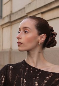 Brązowy sweter dziany z teksturowanym, otwartym wzorem. Włosy związane satynową brązową wstążką i ozdobione złotymi kolczykami w kształcie koła. Starannie ułożony kok.