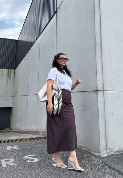Camiseta blanca, falda midi marrón, sandalias transparentes con tacones en bloque. Bolso gris y bufanda estampada como accesorios. Fondo de pared de hormigón.