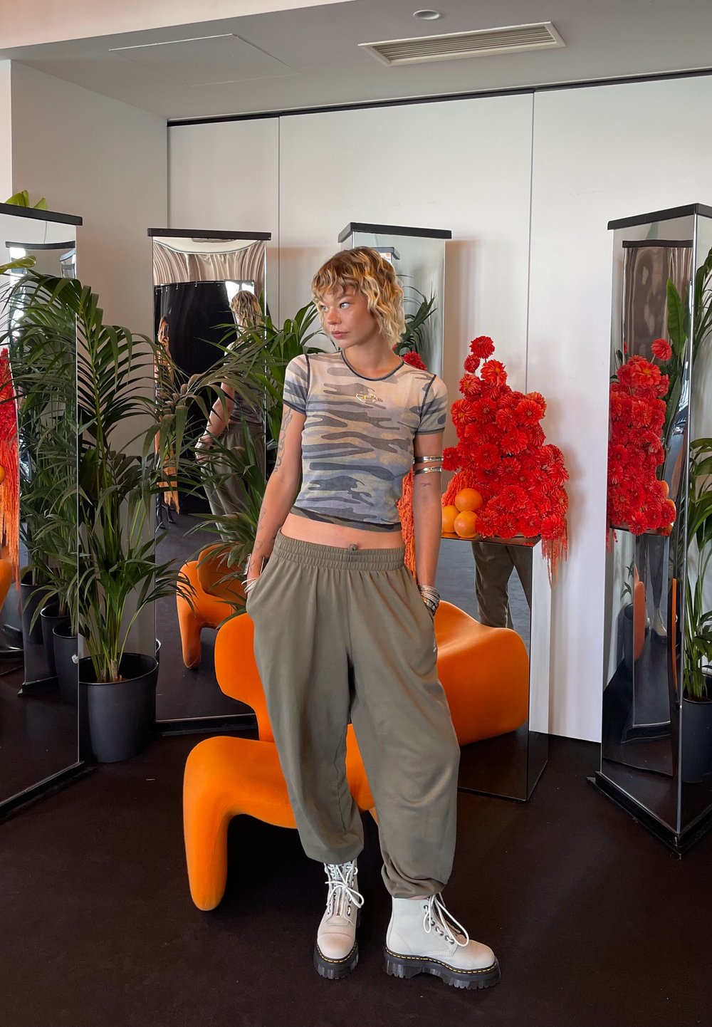 Top corto de camuflaje gris, pantalones holgados verdes oliva y botas de combate blancas con suelas negras. Silla naranja y decoraciones florales rojas en el fondo.