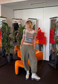 Top corto de camuflaje gris, pantalones holgados verdes oliva y botas de combate blancas con suelas negras. Silla naranja y decoraciones florales rojas en el fondo.