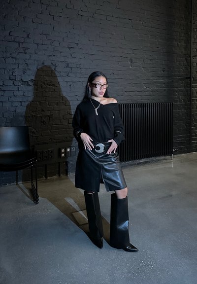 Jersey negro oversize, falda de cuero negro brillante y botas negras hasta la rodilla. Los accesorios incluyen pendientes largos y una gran hebilla de cinturón.