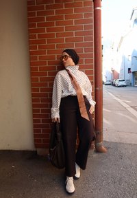 Mujer llevando un pañuelo negro, blusa de lunares con corbata marrón, pantalones negros, zapatillas blancas y gafas de sol, apoyada contra una pared de ladrillo rojo.