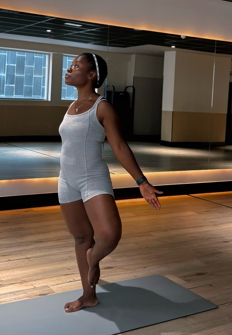 Femme en short de sport gris, en équilibre sur un pied dans une posture de yoga à l'intérieur d'un studio avec un sol en bois et un mur miroir.