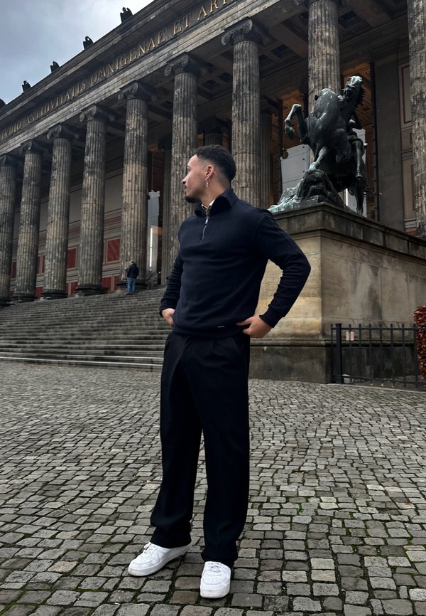 Sudadera negra con cremayera, pantalones negros y zapatillas blancas. Está de pie en unos escalones de piedra en frente de un edificio histórico con columnas.