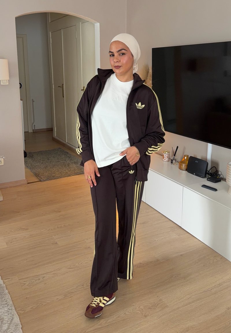 Жена, облечена в тъмен спортен екип на Adidas със жълти ивици и бяла кърпа на главата, стои в модерен хол с телевизор и дървен под.