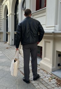 Czarna skórzana kurtka, szare spodnie, trzymając jasną torbę na ramię. Gładka tekstura, luźny krój i minimalistyczny design.