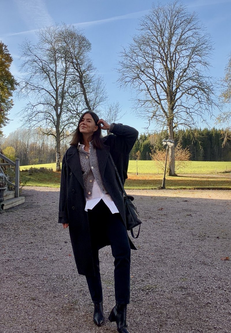 Zwarte oversized jas over een lichtgrijze gebreide top, witte blouse eronder, zwarte broek en enkellaarsjes. Achtergrond omvat bomen en een open veld.