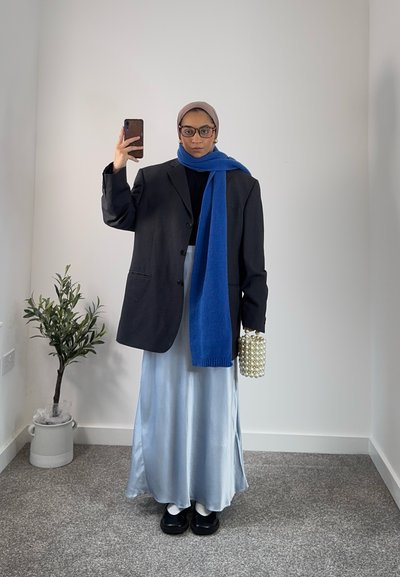 Blazer negro de gran tamaño, bufanda azul, top negro, falda maxi de satén plateado, zapatos negros y bolso con acentos de perlas, contra un fondo neutro.
