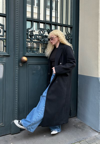 Abrigo negro de lana con diseño de doble botonadura, llevado sobre una camiseta negra y unos jeans de pierna ancha de color azul claro, combinado con zapatillas negras y blancas.