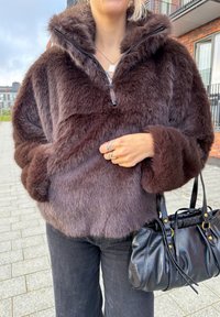 Jachetă maro din blană sintetică cu fermoar frontal și glugă, prevăzută cu mâneci largi și textură moale. Asortată cu o geantă de mână din piele neagră.