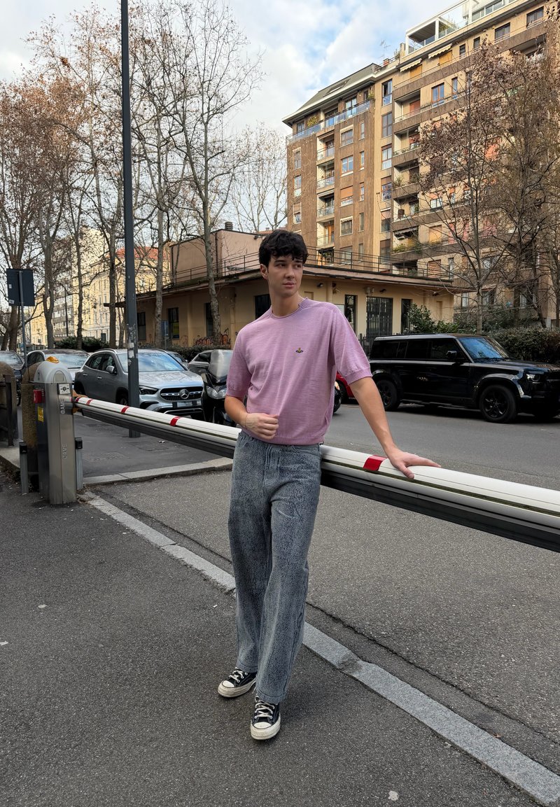 Jeune personne portant un t-shirt violet et un jean large appuyée sur une barrière de parking baissée dans une rue urbaine avec des voitures garées et des arbres dépouillés.